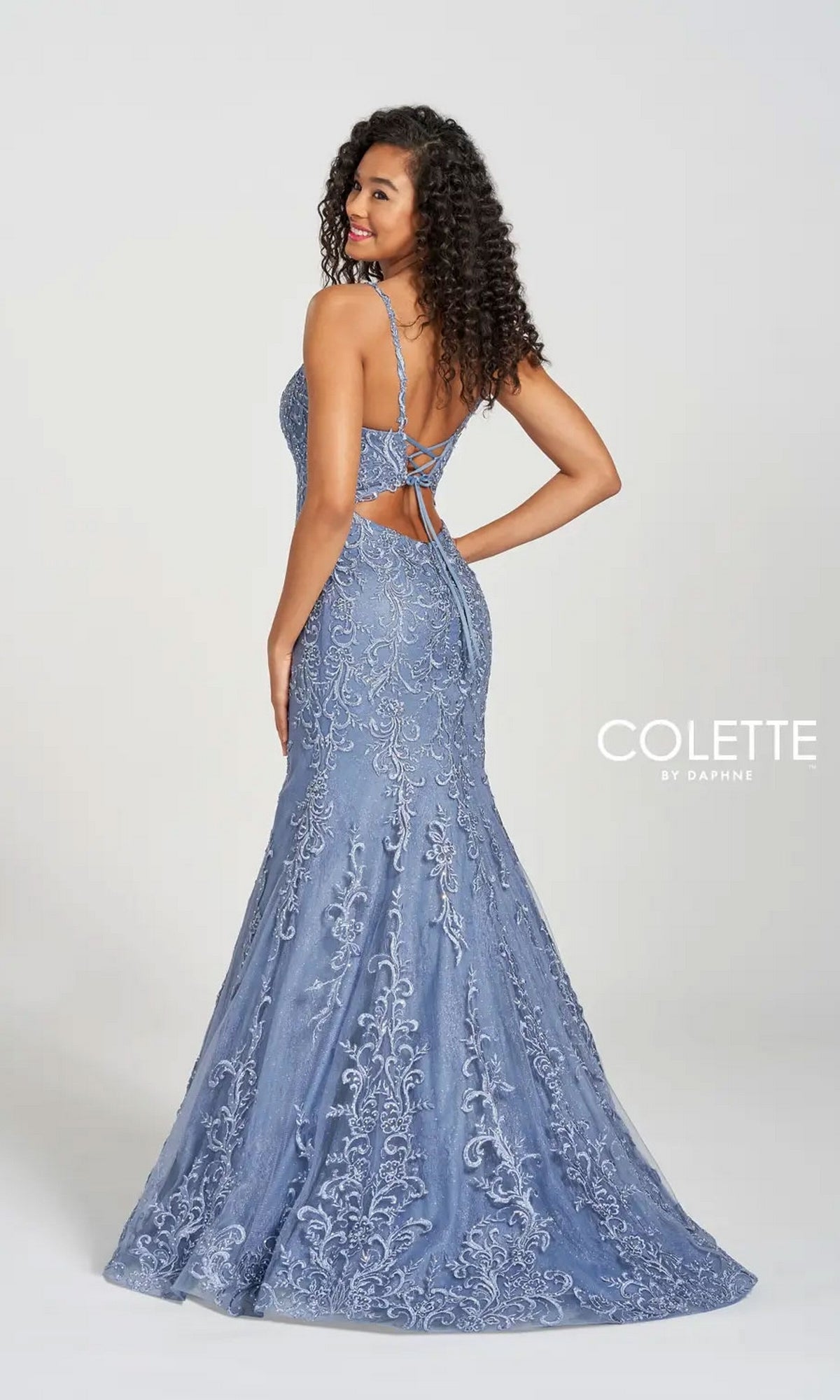 A woman smiles over her shoulder in the Long Prom Dress CL12220 by Colette, a blue formal gown with lace details, thin straps, a v-neck open back, and classic mermaid silhouette. The